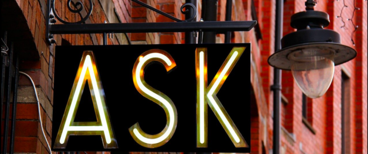 A black sign hanging from a red brick building with the word Ask in shiny gold letters. There is also light hanging next to the sign.
