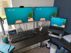 A photo of Dan's desk with a All Things IC desktop background on three computer screens and an All Things IC notebook and pen on the black desk
