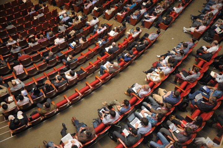 A photo of an auditorium, with red seats and half of them filled with people. The photo has be taken from above.