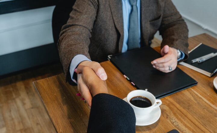 Two people shaking hands over a wooden office table with a cup of coffee in between them
